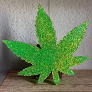 May include: A green glitter cannabis leaf decoration. The leaf has a textured surface with yellow glitter accents. The decoration is placed on a wooden surface with a neutral background.
