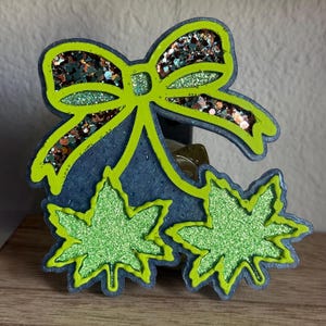 May include: A decorative resin piece with a neon green bow and two cannabis leaf shapes. The bow is filled with glitter in shades of green, brown, and gold. The leaves are filled with green glitter and outlined in neon green.