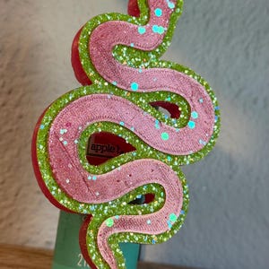 May include: A snake-shaped decorative object featuring a layered design with a metallic pink top layer and a bright green glittery base layer. The object has a textured surface and is positioned vertically, displaying a coiled, winding body shape.