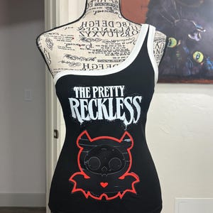May include: A black ribbed tank top with white trim featuring the text THE PRETTY RECKLESS in white lettering. Below the text is a graphic of a black bat-like creature with large eyes and a small red heart, outlined in a bright red border.