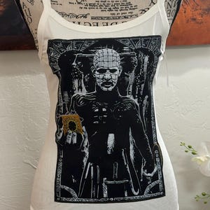 May include: White ribbed tank top featuring a black and white graphic of a character with a grid-like pattern on the head. The graphic is framed by an ornate border. The tank top has thin straps and a scoop neckline.