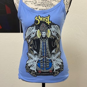 Pode incluir: Top de alças canelado azul claro com um gráfico de uma figura em vestes pretas, rodeada por anjos, com a palavra "Ghost" acima e "Papa Emeritus IV" abaixo. O top tem alças finas e uma silhueta ajustada.