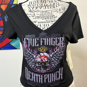 Pode incluir: Camiseta preta com decote em V com um gráfico com uma caveira com asas, uma coroa e o texto "FIVE FINGER DEATH PUNCH". O design incorpora elementos rosa, roxos e brancos. A camisa tem mangas curtas.