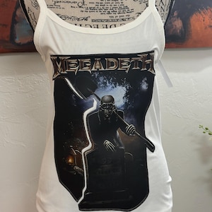 Könnte beinhalten: Weißes geripptes Tanktop mit einem Aufdruck einer Sensenmann-Figur mit Grabstein und dem Bandnamen "MEGADETH". Das Top hat dünne Träger und einen Rundhalsausschnitt.