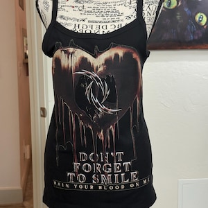 May include: A black tank top featuring a large, dark, dripping heart graphic with a metallic, spiked circular emblem in the center. Below the heart, the text reads DON'T FORGET TO SMILE and RAIN YOUR BLOOD ON ME in a bold, white, serif font.