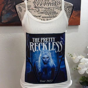 May include: White ribbed tank top featuring a graphic of "THE PRETTY RECKLESS" with a woman's image and "Tour 2022" text. The tank top is displayed on a mannequin.