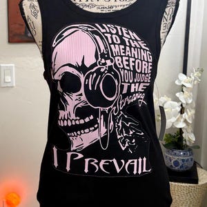 May include: Black tank top with a pink skull graphic wearing headphones. The text on the shirt reads "LISTEN TO THE MEANING BEFORE YOU JUDGE THE SCREAMING" and "I PREVAIL".