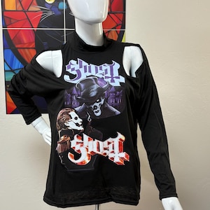 Peut inclure: T-shirt noir à manches longues avec découpes aux épaules. Le t-shirt présente un logo de groupe et des images en blanc, rouge et violet. Le t-shirt est présenté sur un mannequin.