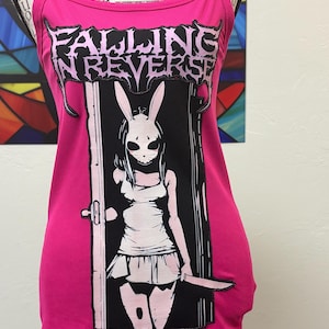 以下が含まれることがあります： 鮮やかなピンク色のタンクトップ。白文字で「FALLING IN REVERSE」と書かれています。ウサギのマスクをつけた人物がナイフを持っているグラフィック。タンクトップはリブ編みです。