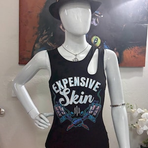 May include: A black tank top with the text "EXPENSIVE SKIN" in white and blue, featuring tattoo machine graphics. The top is displayed on a mannequin, paired with a black hat adorned with embellishments and silver jewelry.