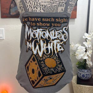 以下が含まれることがあります： 「We have such sights to show you」と「Motionless in White」の文字がスタイリッシュなフォントでプリントされたグレーのタンクトップ。黒と金のキューブのデザインが特徴です。タンクトップは細いストラップが付いています。