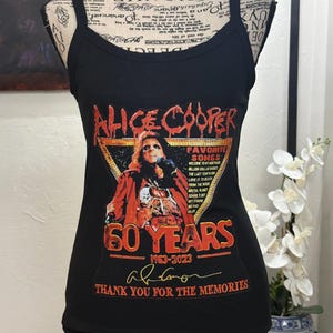 May include: Black tank top featuring a graphic of Alice Cooper with the text "60 Years 1963-2023" and "Thank you for the memories." The design includes a portrait of Alice Cooper and a list of song titles.