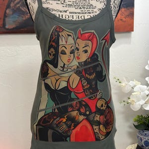 Puede incluir: Camiseta sin mangas verde oliva con un diseño de tatuaje de dos mujeres abrazándose. Una está vestida de monja, la otra de diablo. El diseño incluye calaveras, rosas y un corazón rojo.