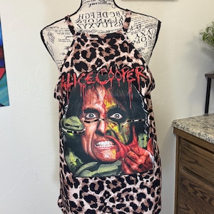 Può includere: Una canotta senza maniche con motivo leopardato marrone e nero. La parte anteriore presenta una grafica di Alice Cooper con un serpente, testo rosso gocciolante e un design a volto diviso. Il capo è esposto su un manichino con stampa a script.