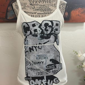 Op de afbeelding: Witte tanktop met een zwart-grijs grafisch ontwerp met de tekst "CBGB" en andere tekst. De tanktop heeft dunne bandjes en een ronde halslijn. De afbeelding bevat de tekst "NY", "315 Bowery", "Since 73" en "CMFUG".