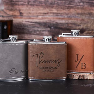 May include: Three personalized leather-wrapped hip flasks with engraved names and dates. The flasks are gray, brown, and brown with a light brown leather finish. The flasks are arranged in a row on a wooden surface.