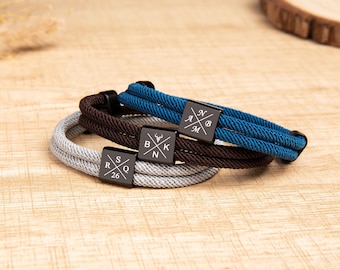 May include: Three layered bracelets featuring blue, brown, and light gray braided cords. Each bracelet has a square black metal charm engraved with a white cross design containing letters and numbers, specifically A, N, B, M, B, K, N, R, S, Q, and 26.
