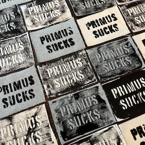 May include: A collection of fabric patches with the phrase "PRIMUS SUCKS" printed in black. The patches are in various shades of gray and white, with a distressed, stencil-like font.