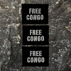 May include: Three black rectangular patches with the words "FREE CONGO" printed in white, stencil-style lettering. The patches are arranged vertically against a textured, dark gray background.