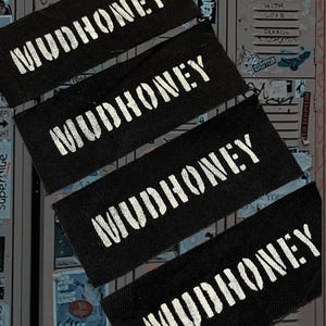 May include: Four black rectangular patches with the word "MUDHONEY" printed in white, stencil-style lettering. The patches are arranged diagonally, slightly overlapping. The background is a collage of stickers and graffiti on a metal surface.