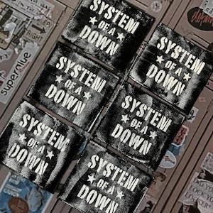 May include: A collection of seven black and white patches. Each patch features the text "SYSTEM OF A DOWN" in a bold, stylized font with a star graphic. The patches are arranged on a surface with various stickers.