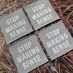 May include: Four square patches with the text "STOP MAKING SENSE" in white stencil font. The patches are a light gray denim material with frayed edges. The background is a bed of brown pine needles.