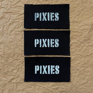 May include: Three black rectangular patches with the word "PIXIES" embroidered in white, stacked vertically. The patches are on a brown, textured surface.