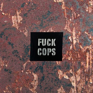 May include: A black square with the words "FUCK COPS" in white, stencil-style lettering. The square is set against a textured background of peeling, rusty red and blue paint.