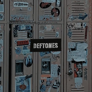 May include: A weathered, brown locker door covered in stickers and graffiti. A black rectangle with the band name "DEFTONES" is centered on the door. The locker is aged and shows signs of wear.