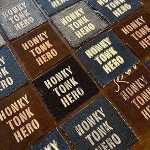 May include: A collection of square fabric patches in various shades of blue, brown, and black. Each patch features the white text "HONKY TONK HERO" in a bold, sans-serif font, suggesting a country music or vintage theme.