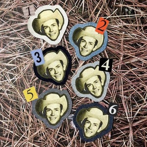May include: Six patches featuring a smiling man wearing a cowboy hat. Each patch has a different colored border and a number from 1 to 6 in the corner. The patches are arranged on a bed of brown and tan pine needles.