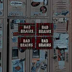 May include: Four rectangular patches with the text "BAD BRAINS" in white, set against a red and black plaid pattern. The patches are displayed against a backdrop of a locker covered in stickers and graffiti.