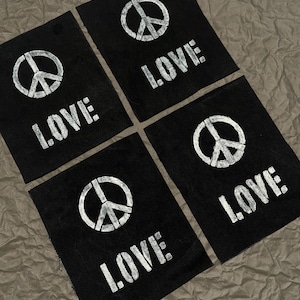 May include: Four black fabric patches with white text and graphics. Each patch features a peace symbol and the word "LOVE" in a bold, sans-serif font. The patches are arranged on a textured, brown surface.