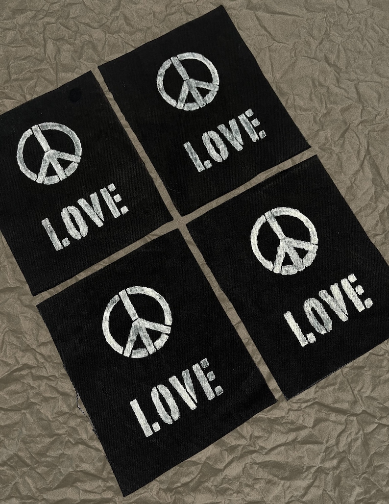 May include: Four black fabric patches with white text and graphics. Each patch features a peace symbol and the word "LOVE" in a bold, sans-serif font. The patches are arranged on a textured, brown surface.