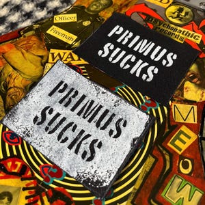 May include: Two black and white iron-on patches with the text "Primus Sucks" in white lettering. The patches are on a colorful background with various patterns and text.