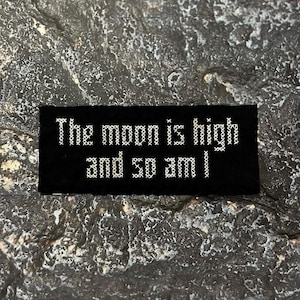 May include: A black rectangular patch with white cross-stitch text that reads, "The moon is high and so am I." The patch is set against a textured, gray stone surface.