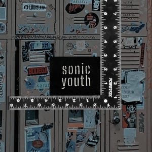 May include: A black square patch with the text "sonic youth" in white lettering is placed over a ruler. The background features a series of lockers covered in stickers and graffiti, creating a vintage aesthetic.