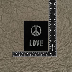 May include: A black square patch featuring a white peace symbol and the word "LOVE" in a stencil font. The patch is placed on a textured, brown surface. A ruler is placed next to the patch.