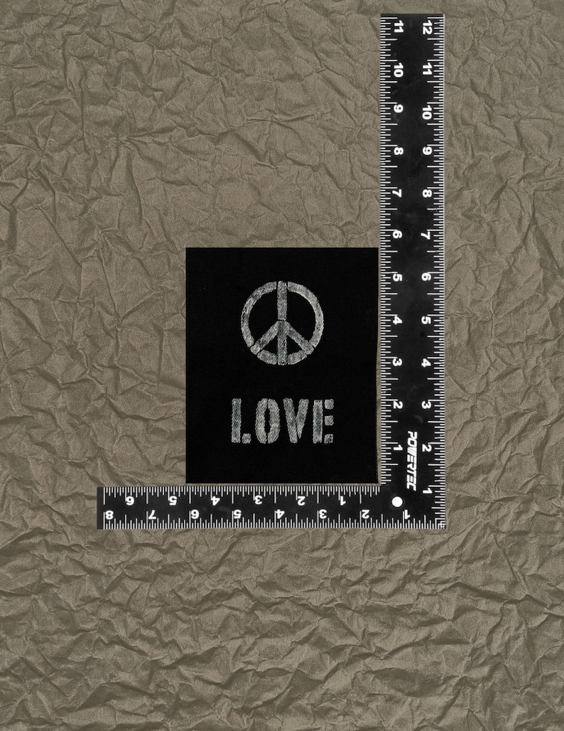 May include: A black square patch featuring a white peace symbol and the word "LOVE" in a stencil font. The patch is placed on a textured, brown surface. A ruler is placed next to the patch.