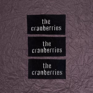 May include: Three rectangular black fabric patches featuring the white embroidered text the cranberries in a lowercase serif font. The patches are arranged vertically, each displaying the same band name in a consistent style and layout.