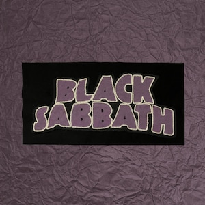 May include: A black rectangle with the words "BLACK SABBATH" in purple letters with a white outline. The background is a textured, dark purple.