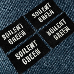 May include: Four black rectangular labels with the text "SOILENT GREEN" in white, arranged on a blue denim background. The labels are evenly spaced and angled, creating a visually appealing pattern. The text is in a bold, sans-serif font.