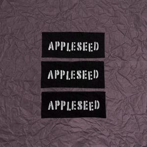 May include: Three rectangular black fabric labels arranged vertically. Each label features the word APPLESEED embroidered in white, sans-serif, stencil-style capital letters. The text is centered on each individual black patch.
