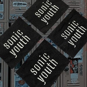 May include: Four black fabric patches with the white text "sonic youth" arranged in a diamond pattern. The background is a locker door covered in graffiti and stickers.