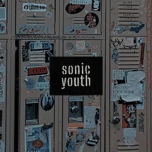 May include: A close-up of a series of lockers covered in stickers and graffiti. A black square with the words "sonic youth" in white lettering is centered in the image. The lockers are a muted brown color.
