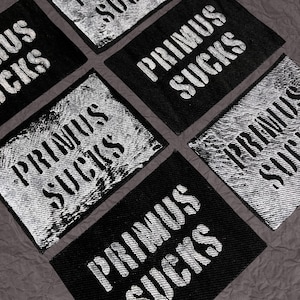 May include: A collection of rectangular fabric patches featuring the text PRIMUS SUCKS in a bold, distressed white font. Some patches have a solid black background, while others display a textured, weathered white finish with black lettering.
