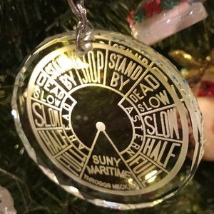 May include: A clear, faceted glass Christmas ornament. The ornament features a circular design with the words "DEAD SLOW," "HALF," and "FULL" in white lettering. The text "SUNY MARITIME THROGGS NECK" is also visible.