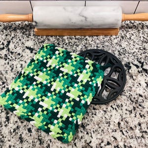 May include: A white marble rolling pin with wooden handles rests on a wooden stand. A green, white, and black woven hot pad sits next to a black cast iron trivet. Kitchen and baking supplies.
