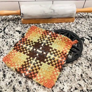 May include: A woven, square hot pad in shades of orange, yellow, brown, and green, resting on a black trivet. A marble rolling pin with wooden handles sits in the background, suggesting a kitchen setting.