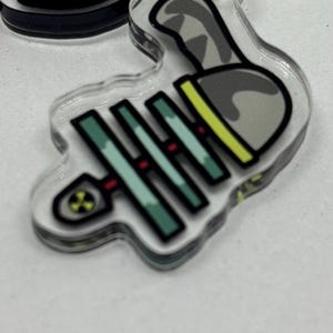 Puede incluir: Un llavero acr&iacute;lico transparente con un dise&ntilde;o de dibujos animados de una espina de pescado y un bolo de bolos. La espina de pescado es de color verde azulado con detalles rojos, y el bolo es gris con detalles amarillos y negros.